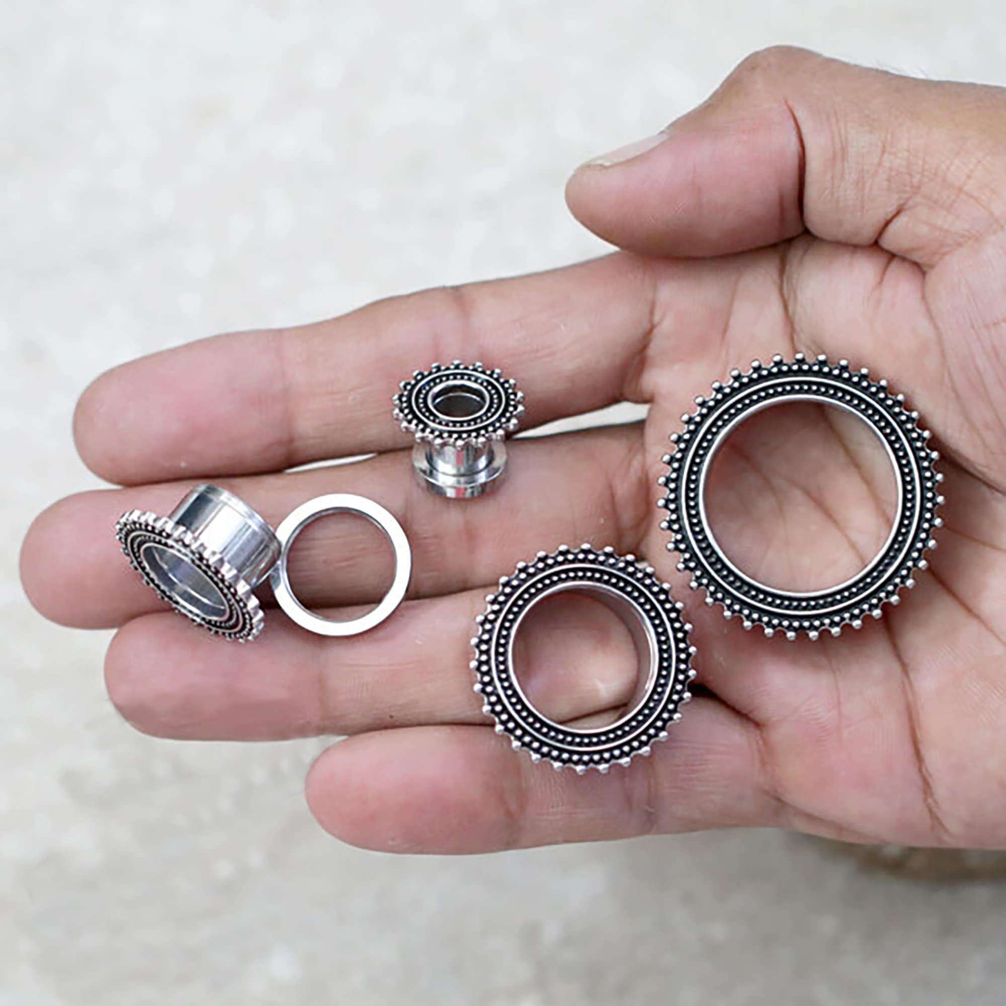 A hand holding several Kaya Movement stainless steel ear gauges in silver and black, featuring intricate beaded designs. The gauges vary in size, with three additional Kaya Movement ear tunnels resting on the palm while one, crafted like a tribal ear tunnel, is held between the thumb and forefinger. The background is a light, neutral surface.
